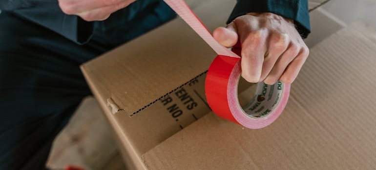A person closing a moving box with red tape 